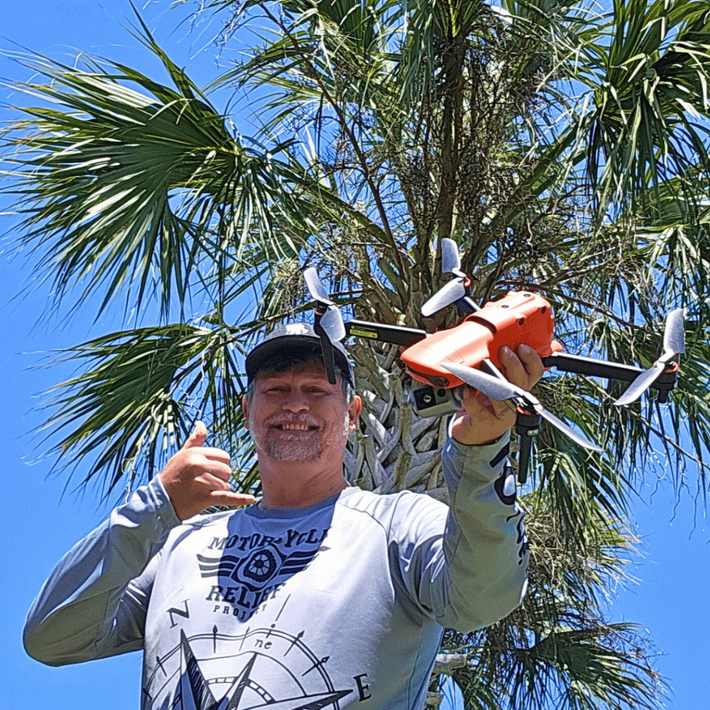 Bob Woodin holding orange drone in front of a palm tree and blue sky, doing hang loose sign